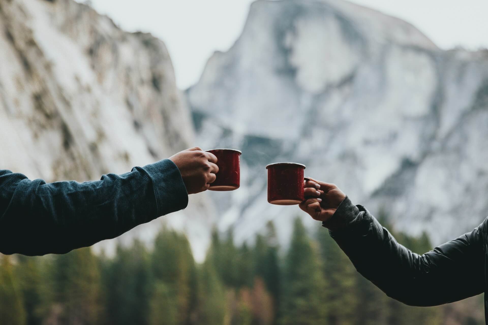 Image shows two different arms raised to clink mugs of hot coffee somewhere outside with mountains.